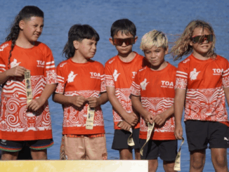A group of young TOA Waka Ama paddlers wearing red team shirts stand together on a podium holding medals, with water visible behind them.