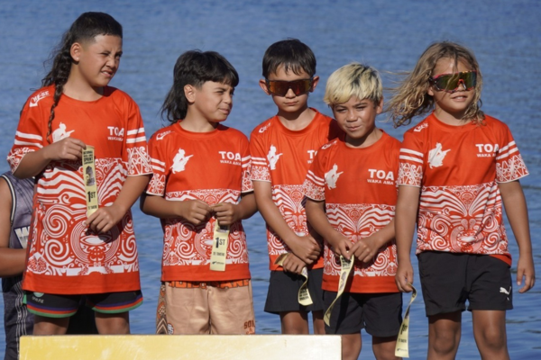 A group of young TOA Waka Ama paddlers wearing red team shirts stand together on a podium holding medals, with water visible behind them.