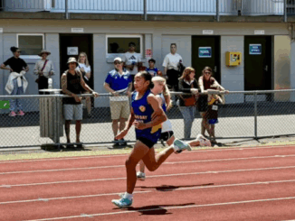 A Titahi Bay junior athlete runs on the track at Newtown Park Stadium, with spectators watching from behind the fence during a junior athletics meet.
