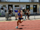 A Titahi Bay junior athlete runs on the track at Newtown Park Stadium, with spectators watching from behind the fence during a junior athletics meet.