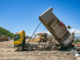 A yellow dump truck tips a large load of rubbish onto a landfill site while heavy machinery operates in the background under a clear blue sky.