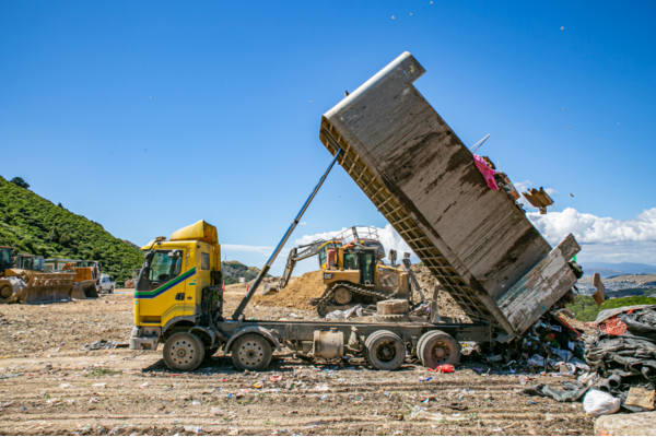A yellow dump truck tips a large load of rubbish onto a landfill site while heavy machinery operates in the background under a clear blue sky.