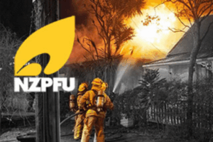 Two firefighters in yellow protective gear spray water on a house fire at night as flames and smoke rise behind trees, with the NZPFU logo displayed in the corner.