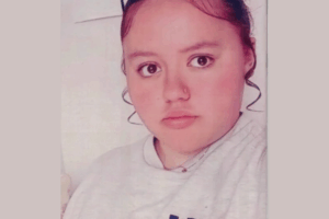 Portrait of a teenage girl with dark hair pulled back, wearing a light grey top and a small nose ring, looking directly at the camera.