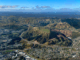 Aerial view of Transmission Gully motorway winding through green hills above Porirua, with suburban neighbourhoods in the foreground and mountains stretching across the horizon under a blue sky.