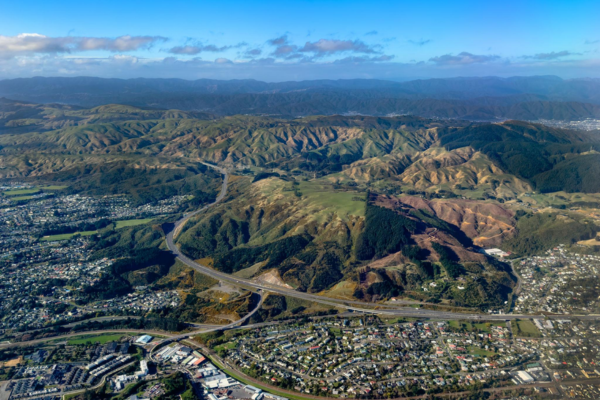 Aerial view of Transmission Gully motorway winding through green hills above Porirua, with suburban neighbourhoods in the foreground and mountains stretching across the horizon under a blue sky.