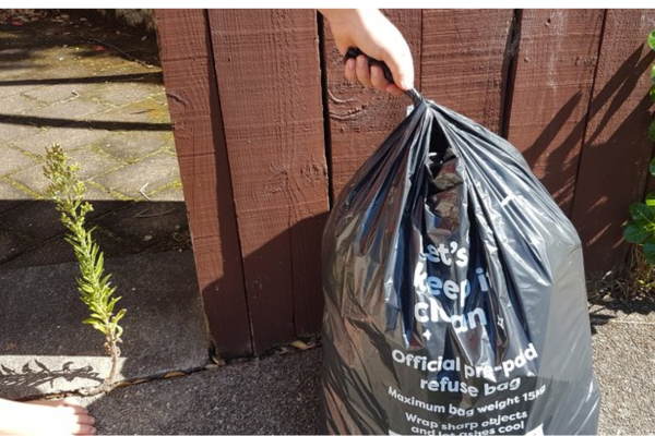 Hand holding a black official Porirua pre-paid rubbish bag beside a wooden fence at a residential kerbside.