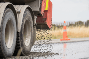 Truck spreads chipseal on wet road surface during highway maintenance works.