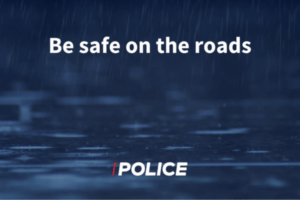 White text reading “Be safe on the roads” over a dark blue background with heavy rain falling on a wet road surface, with a Police logo at the bottom.
