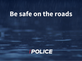 White text reading “Be safe on the roads” over a dark blue background with heavy rain falling on a wet road surface, with a Police logo at the bottom.