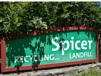 Green roadside sign reading “Spicer Recycling… Landfill” mounted on a low stone wall with trees behind.