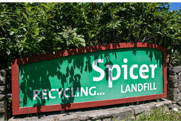 Green roadside sign reading “Spicer Recycling… Landfill” mounted on a low stone wall with trees behind.