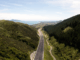 Aerial view of Transmission Gully motorway winding through green hills toward the Kāpiti Coast, with the sea visible in the distance.
