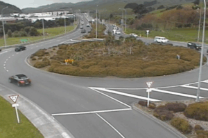 Traffic moves through a large multi-lane roundabout with cars entering and exiting from several directions, surrounded by low shrubs and rolling hills.