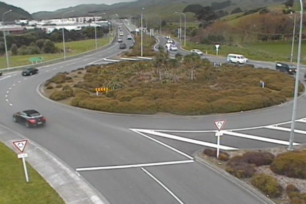 Traffic moves through a large multi-lane roundabout with cars entering and exiting from several directions, surrounded by low shrubs and rolling hills.