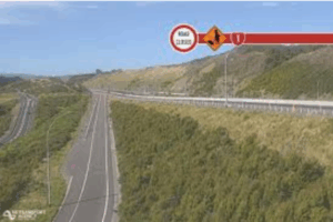 A stretch of Transmission Gully motorway at Waitangirua winds through green hills, with electronic road signs indicating a temporary 50km/h speed limit and lane closure ahead.