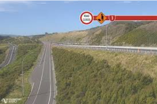 A stretch of Transmission Gully motorway at Waitangirua winds through green hills, with electronic road signs indicating a temporary 50km/h speed limit and lane closure ahead.