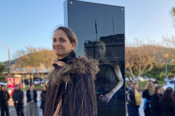 Artist Kohai Grace stands beside a tall glass lightbox artwork during the Te Manawa lightbox unveiling in Porirua in 2022, with people gathered in the background.