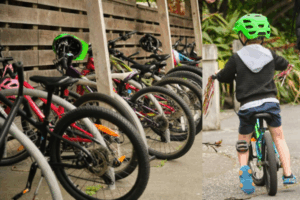 A row of children’s bicycles parked under a wooden shelter at school, alongside a young child wearing a bright green helmet riding a small bike along a footpath.