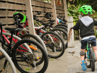 A row of children’s bicycles parked under a wooden shelter at school, alongside a young child wearing a bright green helmet riding a small bike along a footpath.