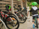 A row of children’s bicycles parked under a wooden shelter at school, alongside a young child wearing a bright green helmet riding a small bike along a footpath.