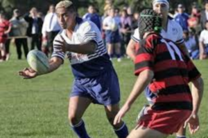 A young rugby player in a blue and white uniform passes the ball during a match, with defenders closing in and spectators watching from the sideline.