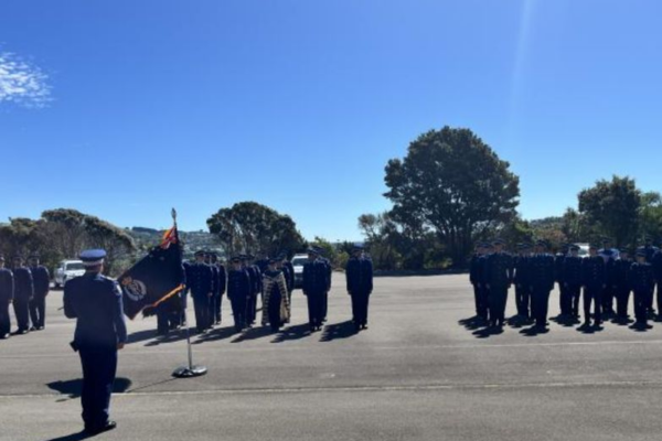 New Zealand Police recruits stand in formation during a graduation parade at the Royal New Zealand Police College in Porirua, with a flag bearer and officer addressing the wing as families watch nearby.