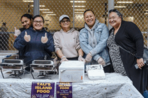 Five women stand behind a food stall at a Pacific night market, smiling and giving thumbs up beside trays of island food.