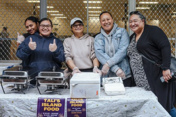 Five women stand behind a food stall at a Pacific night market, smiling and giving thumbs up beside trays of island food.