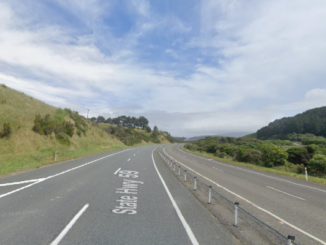 A divided section of State Highway 58 in Porirua, with two lanes in each direction separated by flexible median posts, curving through green hills under a partly cloudy sky.