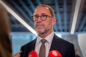 Wellington Mayor Andrew Little speaking to reporters indoors, wearing a suit and glasses, with microphones visible in the foreground.