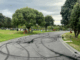 Black burnout tyre marks cover a curved cemetery driveway at Whenua Tapu Cemetery in Porirua, with headstones, green lawns, and trees visible on both sides under a cloudy sky.