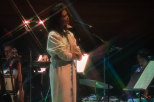 Bic Runga stands at a microphone with hands clasped, singing softly on stage, with orchestral musicians and instruments visible around her under warm, low lighting.