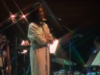 Bic Runga stands at a microphone with hands clasped, singing softly on stage, with orchestral musicians and instruments visible around her under warm, low lighting.