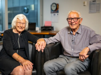 An elderly couple sits side by side in armchairs, smiling at the camera in an office setting.
