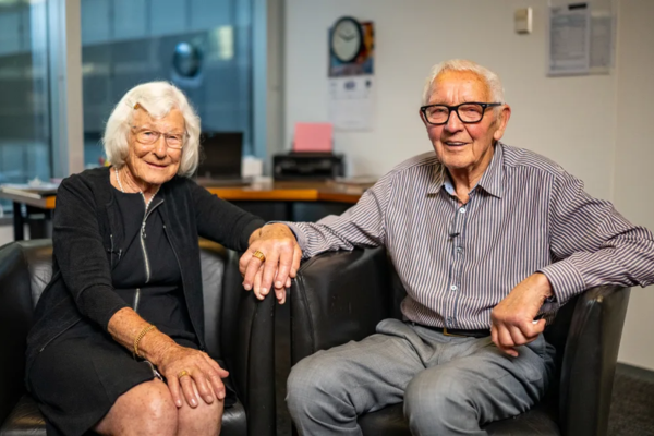 An elderly couple sits side by side in armchairs, smiling at the camera in an office setting.