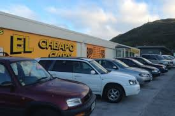 Close-up of a rough asphalt surface beside a concrete kerb, with small stones scattered and shadows from nearby foliage cast across the ground.