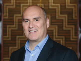 Helmut Modlik wearing a dark blazer and light blue shirt, smiling, standing in front of a patterned wooden wall.