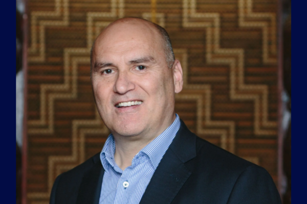 Helmut Modlik wearing a dark blazer and light blue shirt, smiling, standing in front of a patterned wooden wall.