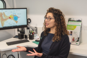 A woman sits in a clinic consulting room, gesturing as she speaks, with a computer screen showing a diagram of the ear behind her.