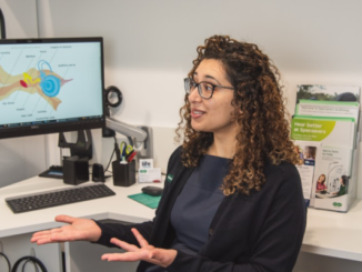 A woman sits in a clinic consulting room, gesturing as she speaks, with a computer screen showing a diagram of the ear behind her.
