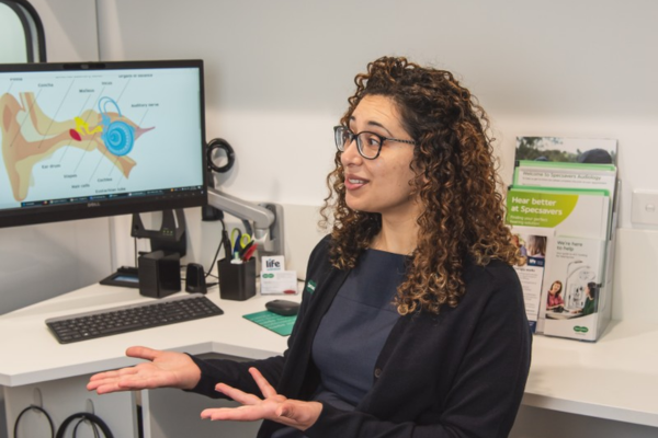 A woman sits in a clinic consulting room, gesturing as she speaks, with a computer screen showing a diagram of the ear behind her.