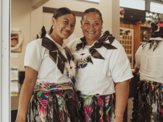 Two Pasifika women stand side by side indoors, smiling at the camera, wearing white tops, woven-style skirts with colourful patterns, and traditional adornments.