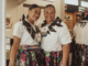 Two Pasifika women stand side by side indoors, smiling at the camera, wearing white tops, woven-style skirts with colourful patterns, and traditional adornments.