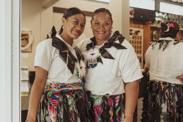Two Pasifika women stand side by side indoors, smiling at the camera, wearing white tops, woven-style skirts with colourful patterns, and traditional adornments.