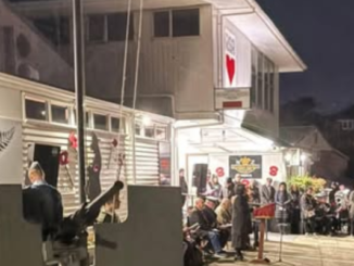 People gather outside the Porirua RSA building at dawn as a service is held, with attendees seated and standing near a flagpole and memorial area.