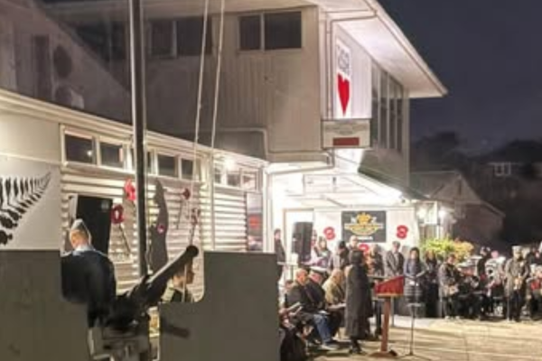 People gather outside the Porirua RSA building at dawn as a service is held, with attendees seated and standing near a flagpole and memorial area.