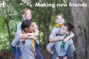 Four children in scout uniforms playing in a wooded area, with two giving piggyback rides, smiling and interacting. Text reads “Making new friends”.
