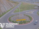 A vehicle drives around a large roundabout beside Transmission Gully motorway, with multiple highway lanes and rocky hillsides visible in the background.
