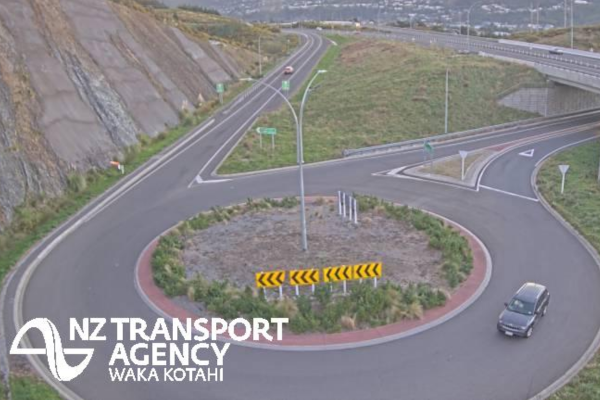 A vehicle drives around a large roundabout beside Transmission Gully motorway, with multiple highway lanes and rocky hillsides visible in the background.